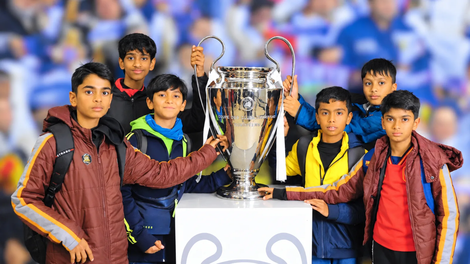 A group of young footballers posing with the UEFA Champions League trophy at a stadium.